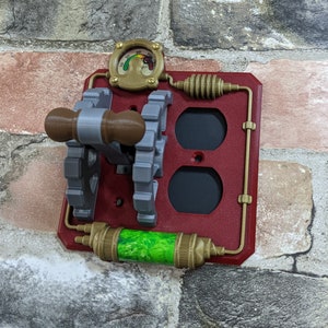 May include: A red and gold steampunk-style outlet cover with a toggle switch, two outlets, and a green gauge. The gauge has a needle pointing to the number 20.