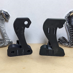May include: Two black, 3D-printed cobra-shaped brackets are shown between two silver cobra emblems. The brackets have a base with two rectangular slots and a hole at the top. The silver emblems are detailed with scales and a menacing expression.