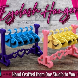 May include: Two eyelash hangers, one purple and one pink, with heart-shaped details. Each hanger has six clear plastic hooks for holding eyelashes. The hangers are hand-crafted and have the text "Eyelash Hanger" and "Hand Crafted from Our Studio to You" on the image.