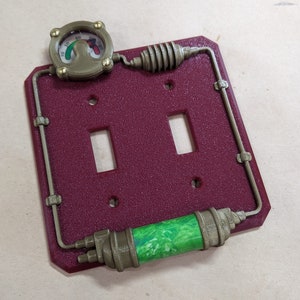 May include: A red and gold light switch cover with a steampunk design. It features a round gauge with a green needle, a pipe-like design, and a green and white cylinder.