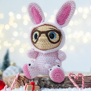 May include: A handmade crochet doll in a pink bunny costume with large ears and a hood. The doll has a tan face, large blue eyes, and wears brown glasses. It is sitting on a wooden surface with Christmas decorations.