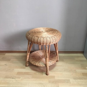 May include: A natural wicker side table with a round top and a lower shelf. The table has four legs and is made of woven wicker. The table is approximately 20 inches in diameter and 20 inches tall.