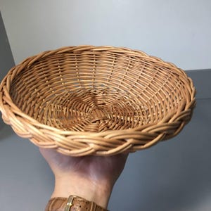 May include: A round, woven wicker basket with a scalloped edge. The basket is light brown and appears to be handmade. The basket is held in a hand, with a brown leather watch strap visible.
