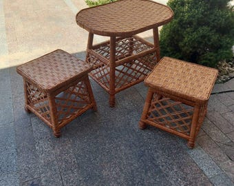 May include: A brown wicker patio set featuring a small oval table and two square stools. The table and stools have a woven design on the tabletop and a lattice pattern on the sides. The set is made of a natural material.