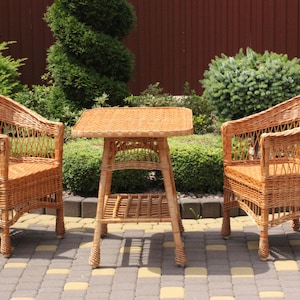 May include: A light brown wicker patio set comprising two armchairs and a small square table. The furniture is made of woven wicker and is set on a brick patio. The chairs have curved backs and armrests, and the table has a lower shelf.