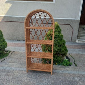 Puede incluir: Una estantería de mimbre marrón claro con cuatro estantes y una parte superior arqueada. Los estantes son rectangulares, y los lados y la parte superior presentan un tejido en forma de diamante. La unidad está hecha de mimbre tejido y se apoya en cuatro patas cortas.