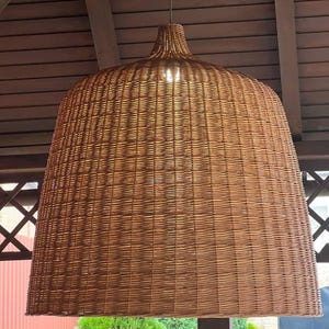 May include: A large, woven rattan pendant light fixture. The light has a bell-shaped shade with a wide base and a narrower top. The fixture is suspended from a white ceiling mount. The light is a warm, natural brown color.