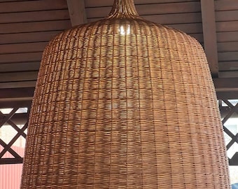 Puede incluir: Una lámpara colgante grande de ratán tejido. La pantalla tiene forma de campana con una base ancha y una parte superior más estrecha. La lámpara está suspendida de un soporte de techo blanco. La luz es de color marrón natural cálido.