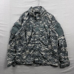 May include: A digital camouflage jacket in grey, black, and white. The jacket has a high collar, multiple pockets, and patches on the sleeves. It is hanging on a metal hanger.