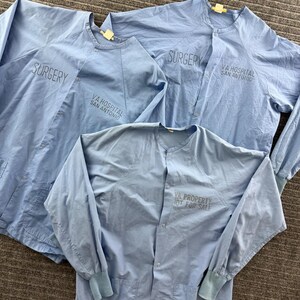 Puede incluir: Tres camisas médicas de manga larga en color azul claro con cierres de botones a presión y puños acanalados. Dos camisas tienen el texto SURGERY y VA HOSPITAL SAN ANTONIO. La tercera tiene el texto VA PROPERTY NOT FOR SALE.