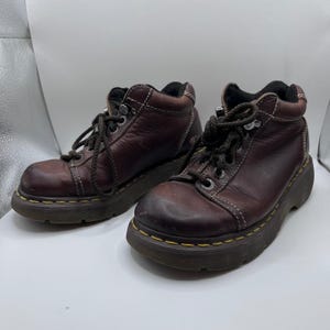 May include: Pair of brown leather ankle boots with laces. The boots have a rounded toe, a thick sole, and white stitching. The soles are outlined with yellow stitching. The boots are on a white surface.