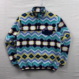 Könnte beinhalten: Buntes Fleece-Pullover mit geometrischem Muster in Blau-, Gelb-, Lila- und Weißtönen. Der Pullover hat einen Stehkragen, einen Druckknopfverschluss und eine kleine Brusttasche. Das Markenlogo ist auf der Brusttasche sichtbar.