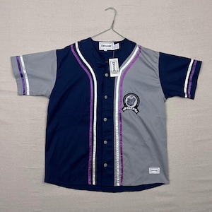 May include: A navy blue and gray baseball jersey with purple and white accents. The jersey has short sleeves and a button-up front. The word "DINAMIC" is printed on the tag and on the jersey. A patch with the word "ROYALS" is on the front.