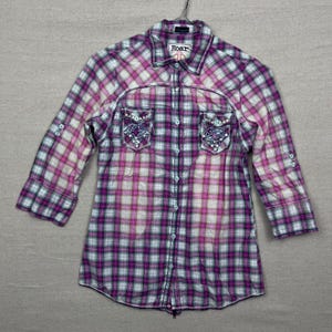 Puede incluir: Camisa a cuadros morada, blanca y azul con mangas tres cuartos. Tiene dos bolsillos delanteros con detalles bordados y la palabra "Roar" en el cuello. La camisa es abotonada.