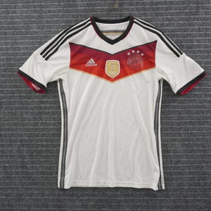May include: A white short-sleeve soccer jersey featuring a V-neck collar, black shoulder stripes, and a red and gold chevron graphic across the chest. The shirt includes an Adidas logo, a FIFA World Cup emblem, and four stars on the front.