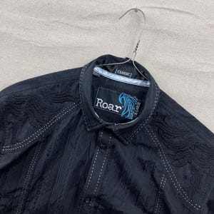 Op de afbeelding: Zwart overhemd met witte stiksels en geborduurde details. Het shirt heeft de merknaam "Roar" op een label en is gemarkeerd als X-Large. Het shirt hangt aan een metalen hanger.