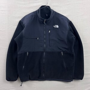 May include: A black and navy blue zip-up fleece jacket. The jacket features a high collar, two side pockets, and a chest pocket. The North Face logo is visible on the upper left chest. The jacket is made of a combination of fleece and nylon materials.