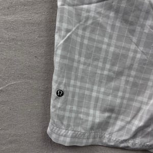 May include: Close-up of a light gray and white plaid fabric. The fabric has a grid pattern and a small black logo on the lower left side. The fabric is folded and appears to be part of a garment.