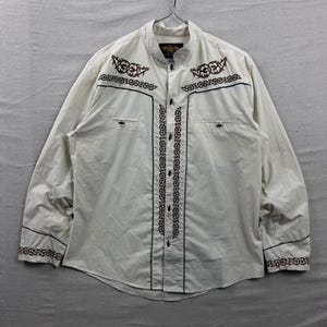 May include: Off-white long-sleeve shirt with intricate brown embroidery and black trim. Features a mandarin collar, button-down front, and two chest pockets. The shirt has decorative patterns on the front, cuffs, and shoulders.