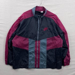 May include: A vintage Nike windbreaker jacket in black, burgundy, and gray. The jacket features a full zipper, a small Nike logo on the chest, and a color-block design. The jacket is hanging on a hanger.