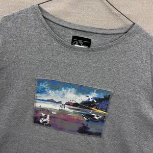 May include: A heather grey t-shirt featuring a rectangular patch on the chest with a printed coastal scene of a beach, ocean, and seagulls. The shirt has a round neckline and a black interior label with white text reading Michael.