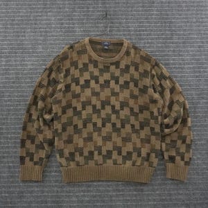 May include: A long-sleeve crew neck sweater featuring a repeating geometric block pattern in shades of tan, brown, and dark olive green. The knit garment has a ribbed collar, cuffs, and hem, with a visible brand label inside the neckline.