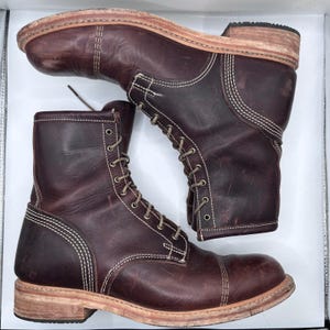 May include: Pair of burgundy leather work boots with cap toes, visible stitching, and light-coloured soles. The boots have a lace-up design with metal eyelets. The boots are set against a white background.