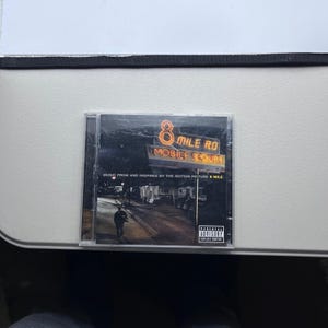 Puede incluir: Una funda de CD para la banda sonora de la película "8 Mile". La portada presenta un letrero de neón que dice "8 Mile Rd Mobile Court". Debajo del letrero está el texto "Music from and inspired by the motion picture 8 Mile". Una advertencia parental está en la esquina inferior derecha.