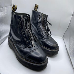 Puede incluir: Par de botas de plataforma de cuero negro con cierre de cremallera frontal y detalles de cordones. Las botas tienen una suela gruesa y el nombre de la marca es visible en la cremallera. Las botas tienen una lengüeta con texto amarillo.