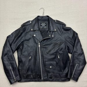 Pode incluir: Jaqueta de motociclista de couro preto com zíperes e ferragens prateadas. Possui gola entalhada, ombreiras e cinto na cintura. A etiqueta diz "ROCKY MOUNTAIN HIDES GENUINE COWHIDE".