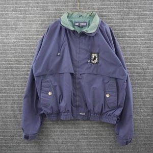 May include: A navy blue zip-up jacket featuring a light green inner collar, a full-length front zipper, and two front pockets with snap closures. A black and white POW-MIA patch is on the chest, and the LA-VING brand label is on the inner collar.
