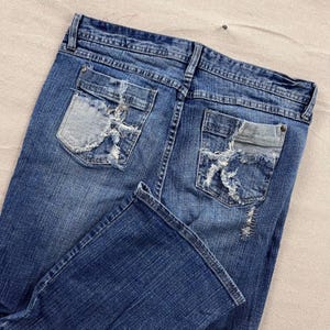 May include: Blue denim jeans with distressed detailing on the back pockets. The jeans have a classic five-pocket design and a faded wash. The back pockets feature patches and ripped accents, adding a vintage, worn look.