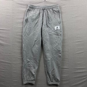 Könnte beinhalten: Hellgraue Jogginghose mit Kordelzugbund und Seitentaschen. Die Hose hat vertikale Nähte auf der Vorderseite und ein kleines weißes quadratisches Patch mit einem schwarzen Logo. Der Stoff scheint ein weiches, bequemes Material zu sein.