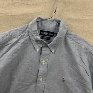 May include: A light gray button-down shirt with a classic collar and long sleeves. The shirt features a small embroidered logo of a polo player on the lower left side. The brand name "Ralph Lauren" is visible on a label near the collar.
