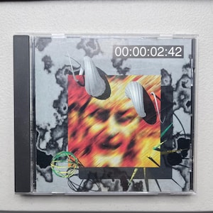 May include: A CD case with an abstract design. The artwork includes a blurred face, red and yellow colours, and white shell-like shapes. The text "00:00:02:42" is displayed at the top. The case has a black spine.