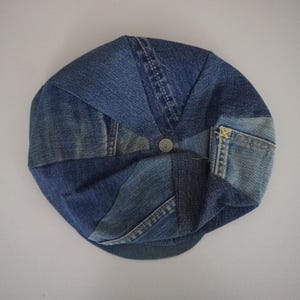May include: A circular flat cap constructed from multiple panels of blue denim fabric in varying shades. The design features visible seams, a central metal button at the crown, and a small pocket detail on the side with yellow stitching accents.
