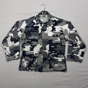 Könnte beinhalten: Eine Tarnjacke in Grau-, Weiß- und Schwarztönen mit Knopfleiste, mehreren Taschen und langen Ärmeln. Die Jacke hängt an einem Metallbügel und zeigt ihr vom Militär inspiriertes Design.