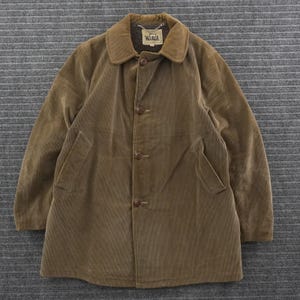 May include: A tan corduroy jacket featuring a fold-over collar, long sleeves, and a front button closure with four visible buttons. The interior label at the neckline displays the Woolrich brand name and a small sheep logo graphic.