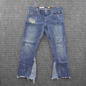 May include: Blue denim jeans featuring a mid-rise waist, a button and zipper fly, and five pockets. The front displays distressed areas, including a leopard print patch on the right thigh and frayed, flared hems with contrasting light blue fabric inserts.