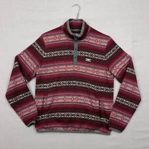 Pode incluir: Um pulôver de lã polar com padrão bordô e cinza, com gola alta e três botões. A camisa de manga comprida apresenta um design geométrico com detalhes em vermelho, preto e branco. Um pequeno logotipo é visível no canto inferior direito.