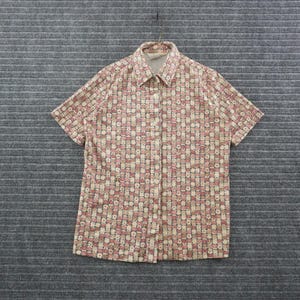 May include: A short-sleeved button-down shirt featuring a repeating checkered pattern in shades of pink, beige, and dark brown. The shirt has a standard collar, a front button placket with light-colored buttons, and a straight hemline.