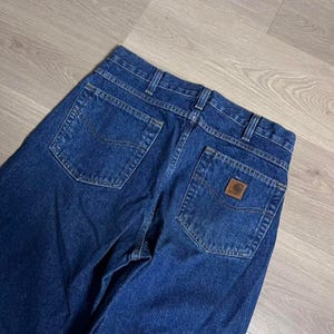 May include: Dark blue denim jeans with a classic five-pocket design. The back pockets have a curved shape, and a brown leather patch with a logo is visible on the back right pocket. The jeans are made of a sturdy denim material.