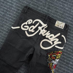 May include: Black denim jeans featuring a large, cream-coloured script logo that reads Ed Hardy across the back pockets. A colourful embroidered tiger face graphic is positioned on the lower right leg area, and a rectangular brand label is attached.