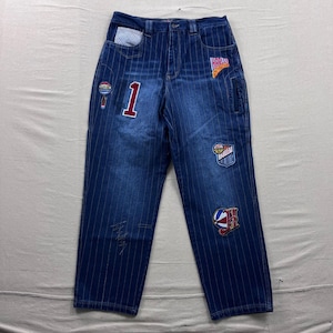 May include: Blue denim jeans with vertical white pinstripes and various patches. Patches include the number "1", a basketball, and the word "Harlem". The jeans have a classic five-pocket design.