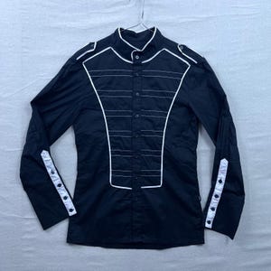 Könnte beinhalten: Schwarzes Langarmhemd mit weißen Zierleisten und Akzenten. Das Hemd hat einen Stehkragen, geknöpfte Manschetten und dekorative weiße Linien auf der Vorderseite. Das Design umfasst Schulterklappen und eine Knopfleiste.