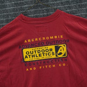 May include: A red t-shirt featuring a rectangular yellow and black patch on the back. The patch reads "SUPPORTING OUTDOOR ATHLETICS FOR OVER 100 YEARS" with an "A" logo. Additional text on the shirt includes "ABERCROMBIE ADVANCED OUTDOOR CONSTRUCTION AND FITCH CO."