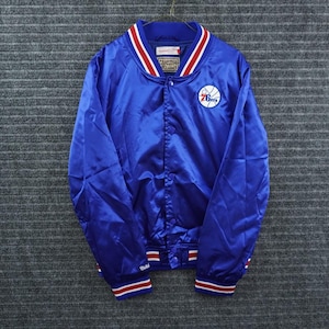 May include: A royal blue satin varsity jacket featuring a white and red striped ribbed collar, cuffs, and hem. The front displays a circular Philadelphia 76ers logo on the left chest, a snap-button closure, and Mitchell & Ness Hardwood Classics labels.