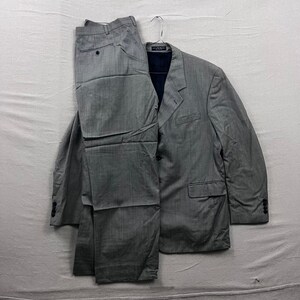 Könnte beinhalten: Ein grauer Anzug, bestehend aus einem Blazer und einer passenden Hose, hängt an einem Metallbügel. Der Anzug hat ein dezentes Streifenmuster. Der Blazer hat ein fallendes Revers, eine Brusttasche und zwei Vordertaschen. Die Hose hat einen Knopf- und Reißverschluss.