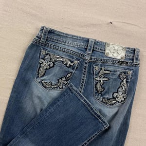 May include: Blue denim jeans with decorative stitching and embellished back pockets. The pockets feature intricate silver and black designs, including fleur-de-lis and cross motifs. A silver label is visible on the back.