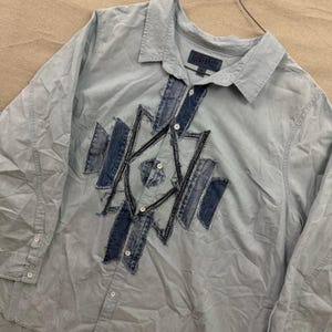 May include: Pale blue button-down shirt with a denim patchwork design. The shirt has a collared neckline, long sleeves, and a button-up front. The denim design is stitched onto the front. The Lucky Brand label is visible.